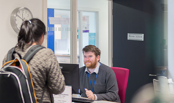 A  staff member smiles up at a student who is in front of his desk