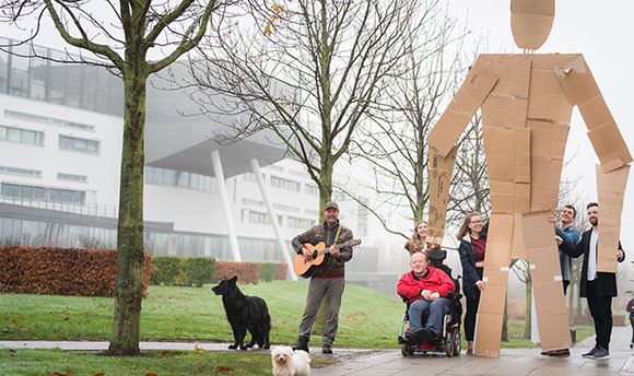 Students outside  holding a giant card board person up whilst a man with a dog plays guitar