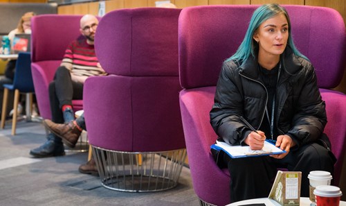 Student with vibrant blue hair sat on purple pod seat in  Atrium